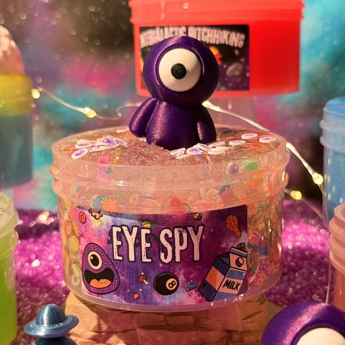 A clear plastic jar filled with colorful glitter and small beads, topped with a purple one-eyed cartoon character. The front label reads &