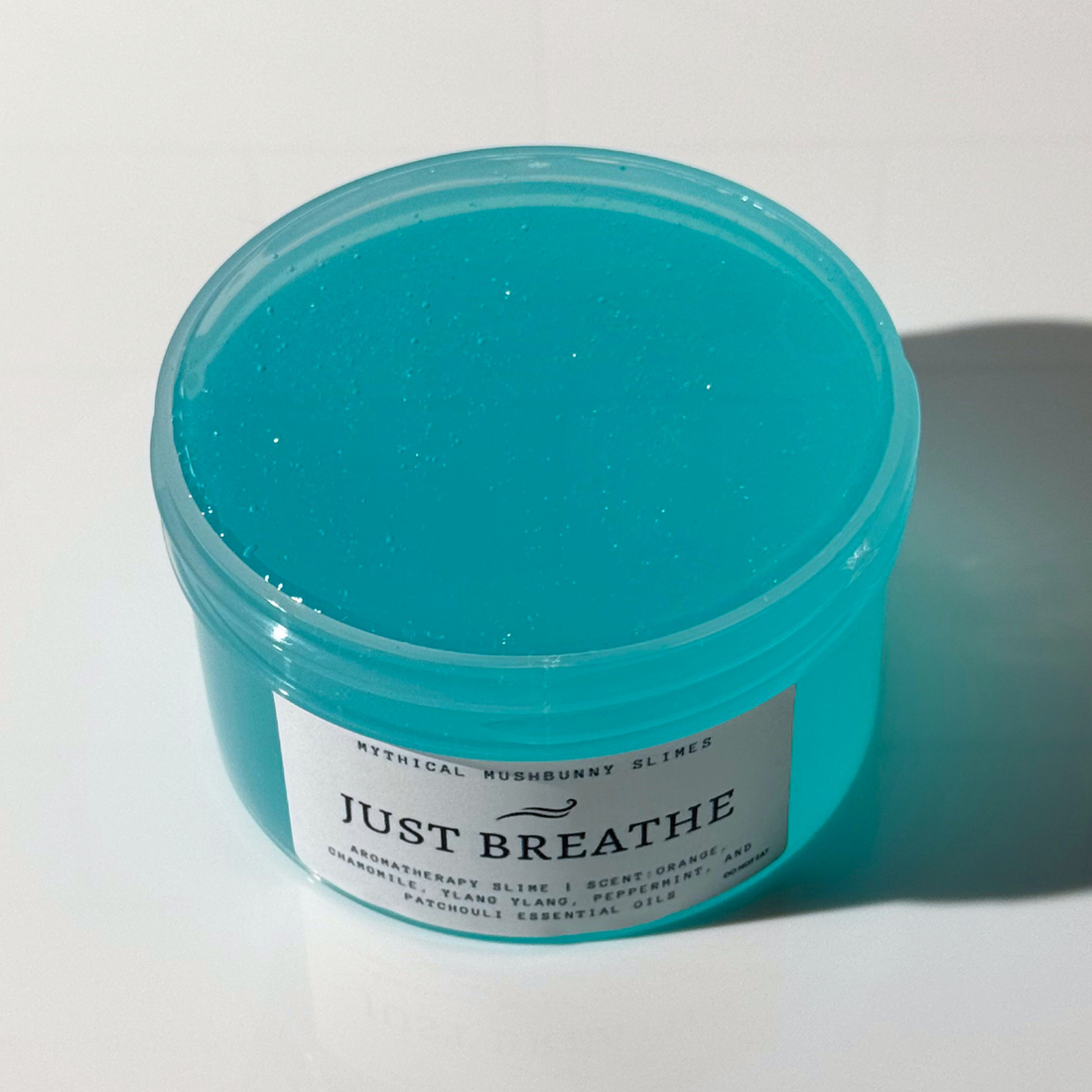 A clear jar of turquoise blue Just Breathe aromatherapy slime with a white label on a clean background.
