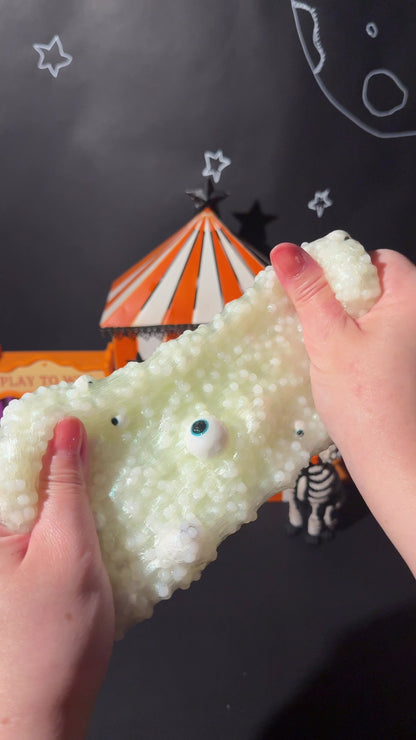 Video demonstration stretching green slush bead Guess How Many Eyes slime packed with eyeball charms in front of a carnival backdrop.