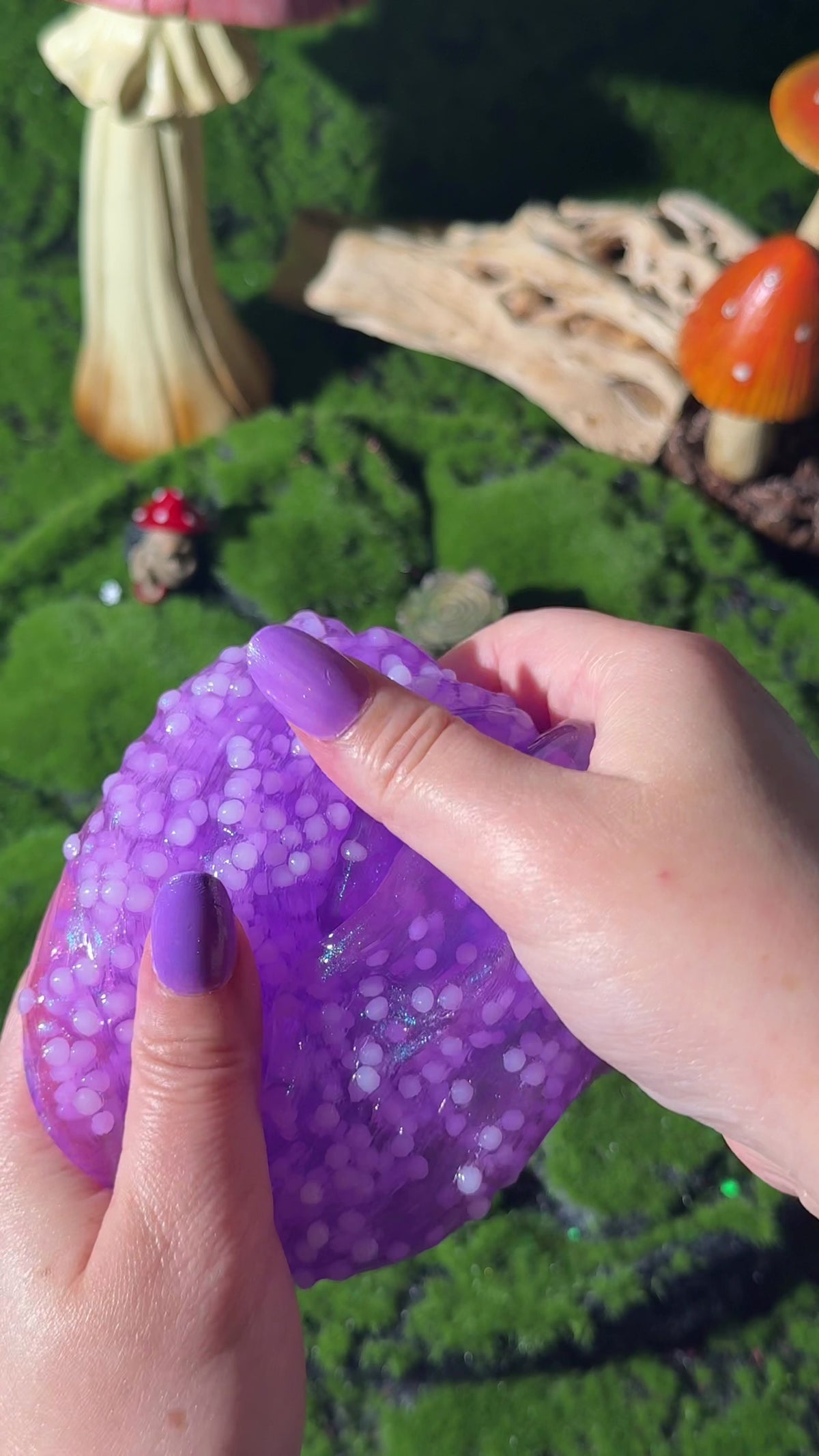 Hands stretching neon purple clear slush slime with iridescent glitter, set outdoors on moss with mushroom decorations.