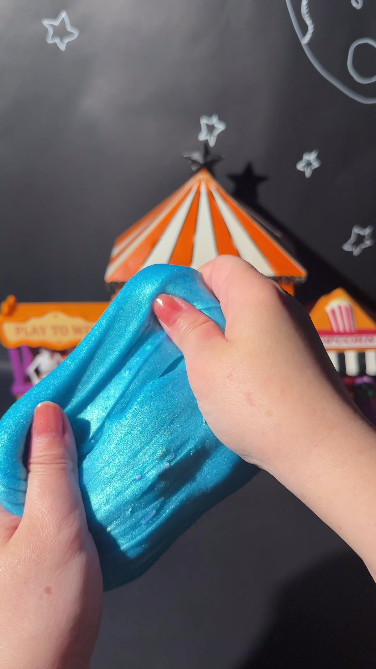 Hands stretching vibrant blue Rancid Refresher jelly slime in front of a carnival circus backdrop