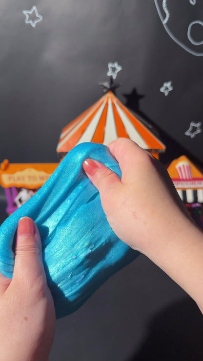 Hands stretching vibrant blue Rancid Refresher jelly slime in front of a carnival circus backdrop